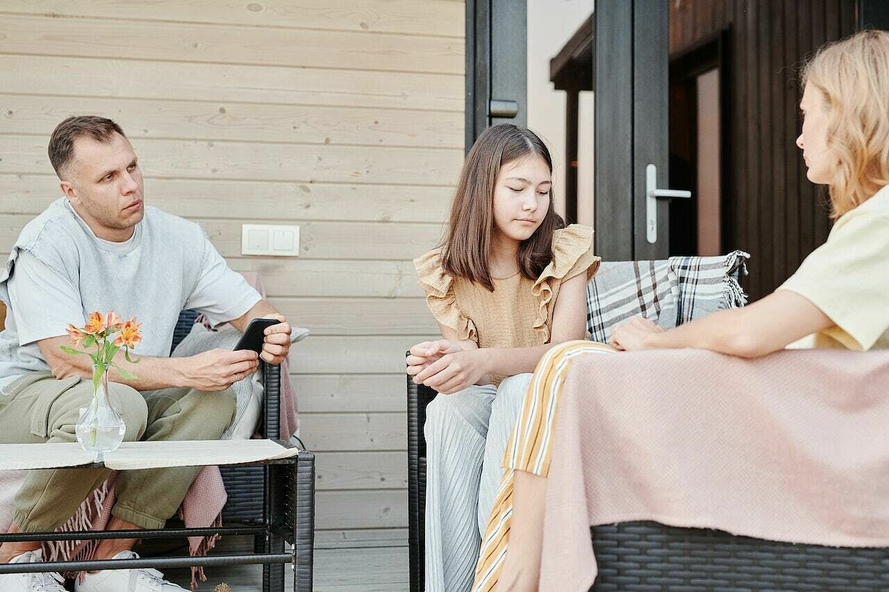 Ein Mann, eine Frau und ein älteres Mädchen sitzen auf Terrassenstühlen einander zugewandt vor einer Holzwand. Die Gesichter sind ernst. Der Mann blickt zur Frau, das Mädchen hat den Blick gesenkt.