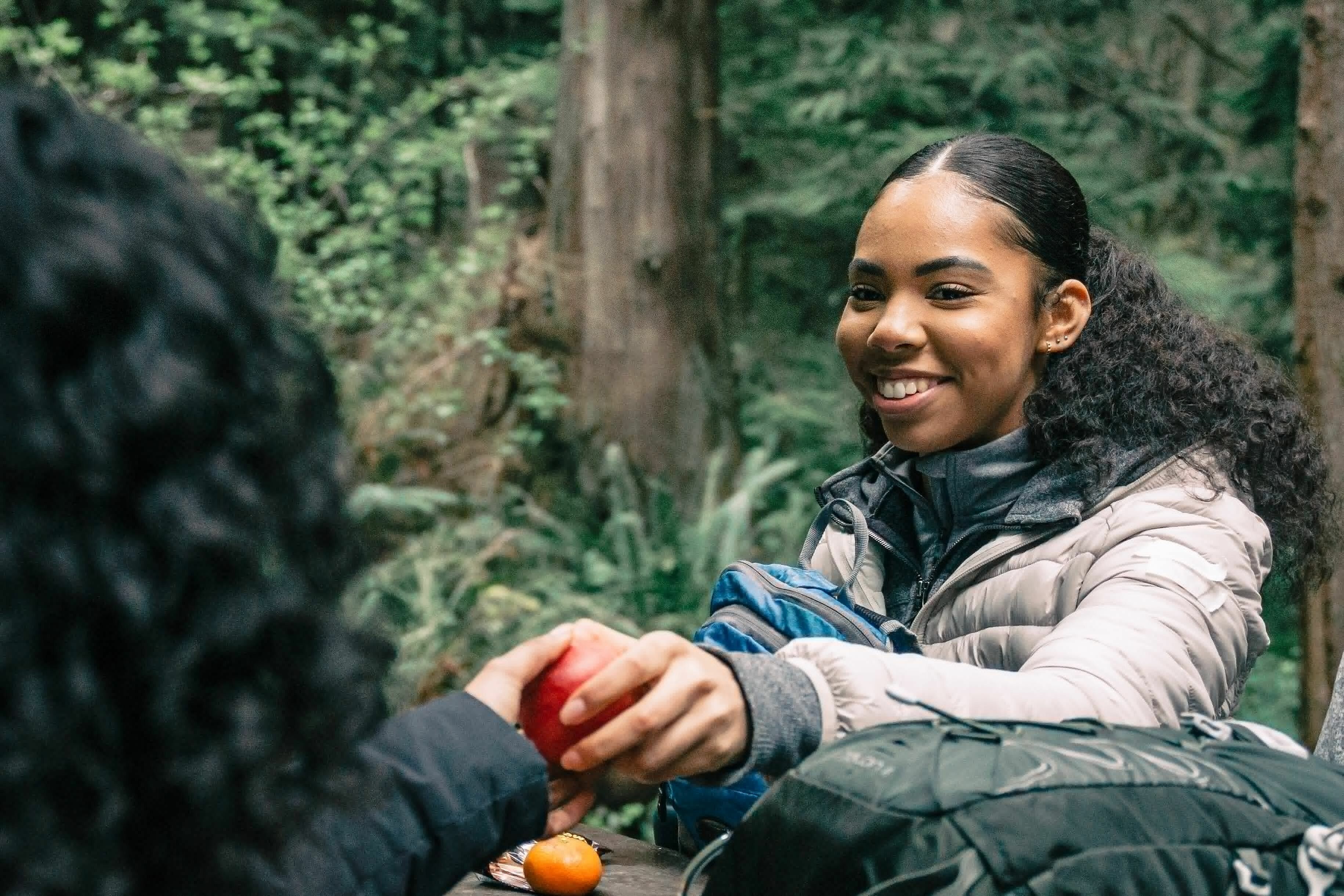 Eine junge Frau mit lockigen, schwarzen Haaren nimmt im Wald grinsend einen Apfel entgegen von einer Person mit schwarzen Locken. 