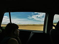 Eine junge Frau schaut aus dem fahrenden Auto, leicht angeleht an der Scheibe. Sie trägt Kabelfreie Kopfhörer. Im Hintergrund sieht man die Landschaft - trockene Felder und Himmel mit Schäfchenwolken.